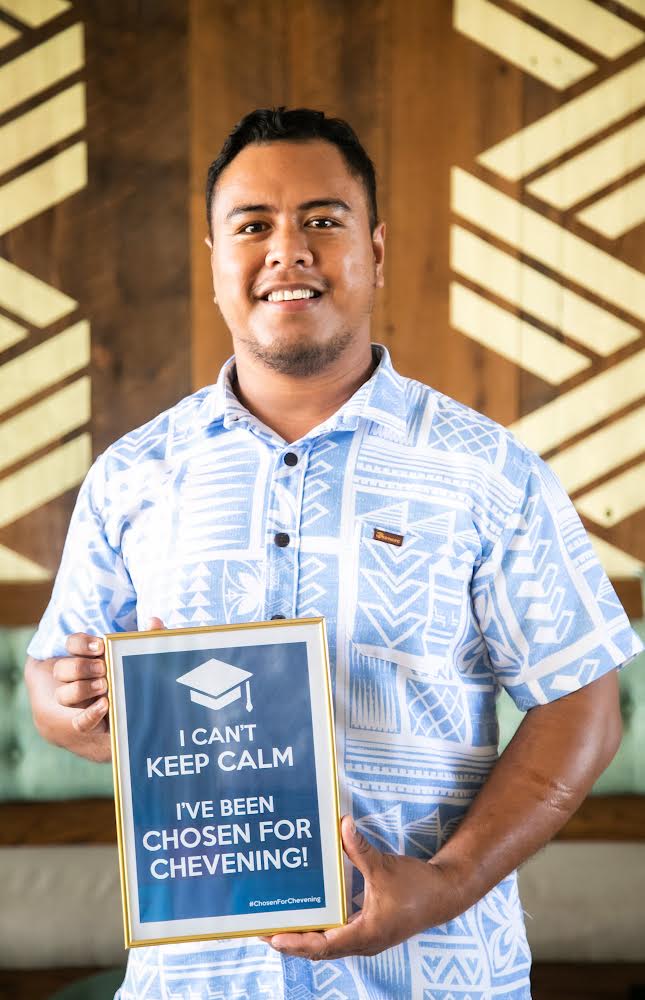 James is wearing a blue patterned shirt and holding a sign that reads 'I can't keep calm I've been chosen for Chevening'