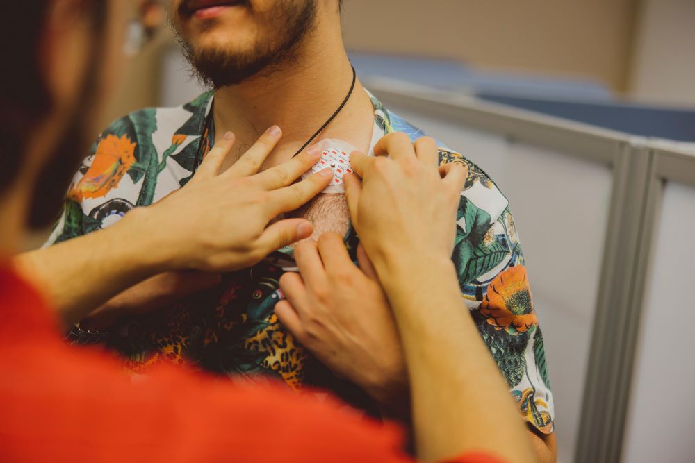 Looking over the shoulder of a person applying a patch to the shoulder of a second figure, mostly out of shot, who is wearing a floral top and has a beard.