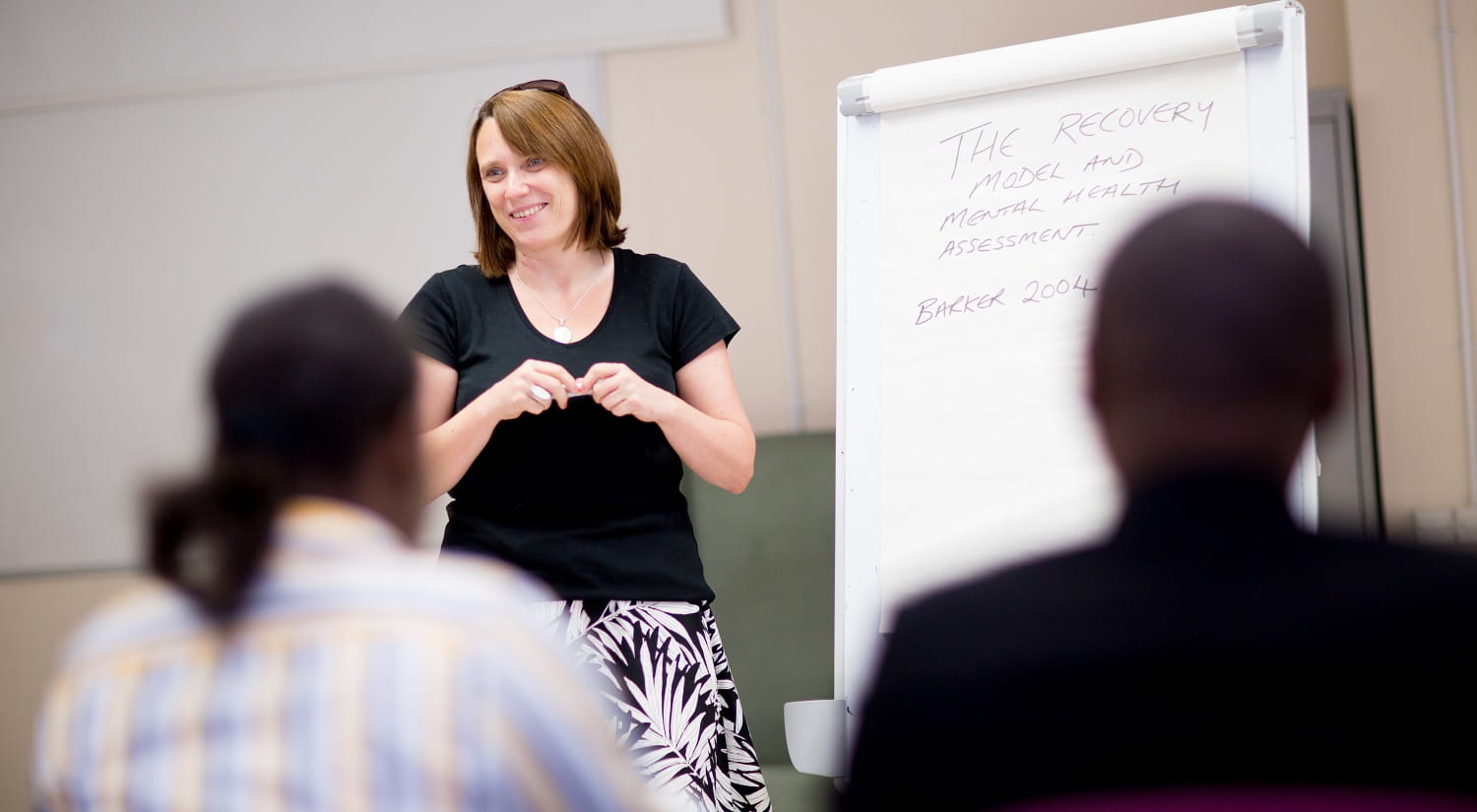 Tessa Wagstaff from the School of Health and Social Care, standing next to a flip chart with "The recovery model and mental health assessment. Barker 2004" written in purple pen. Two students with their backs to the camera are sitting in the foreground.