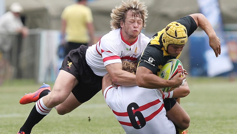 Two rugby players in white shirts with red lines, tackling another player in a black and yellow shirt and a yellow cap on his head, who is holding the rugby ball tucked under his arm.